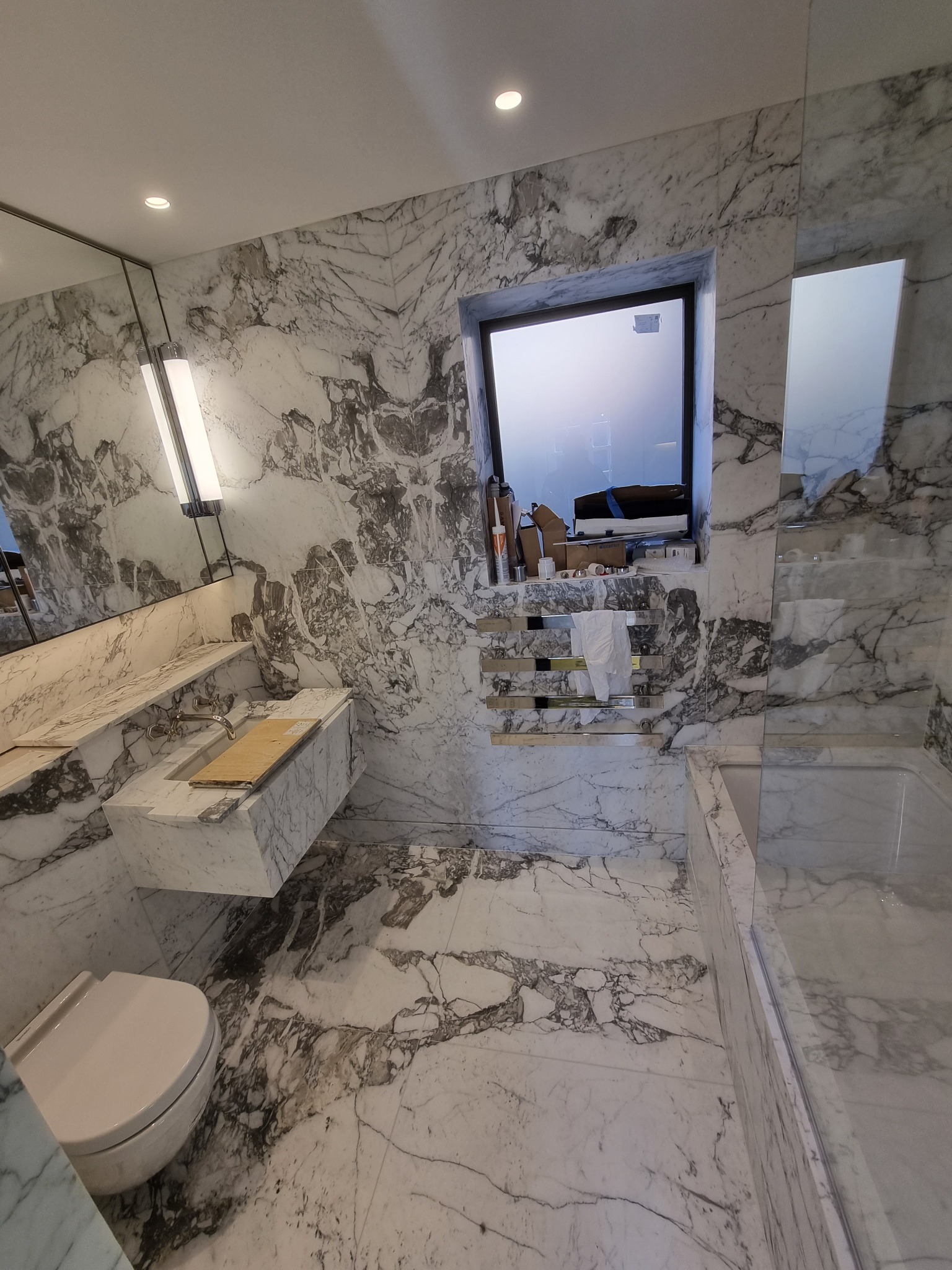 Nowoczesna łazienka wykończona białym marmurem, z podwieszaną umywalką, toaletą i wanną za szklaną ścianką; widoczne materiały i narzędzia przy oknie sugerują trwające prace.