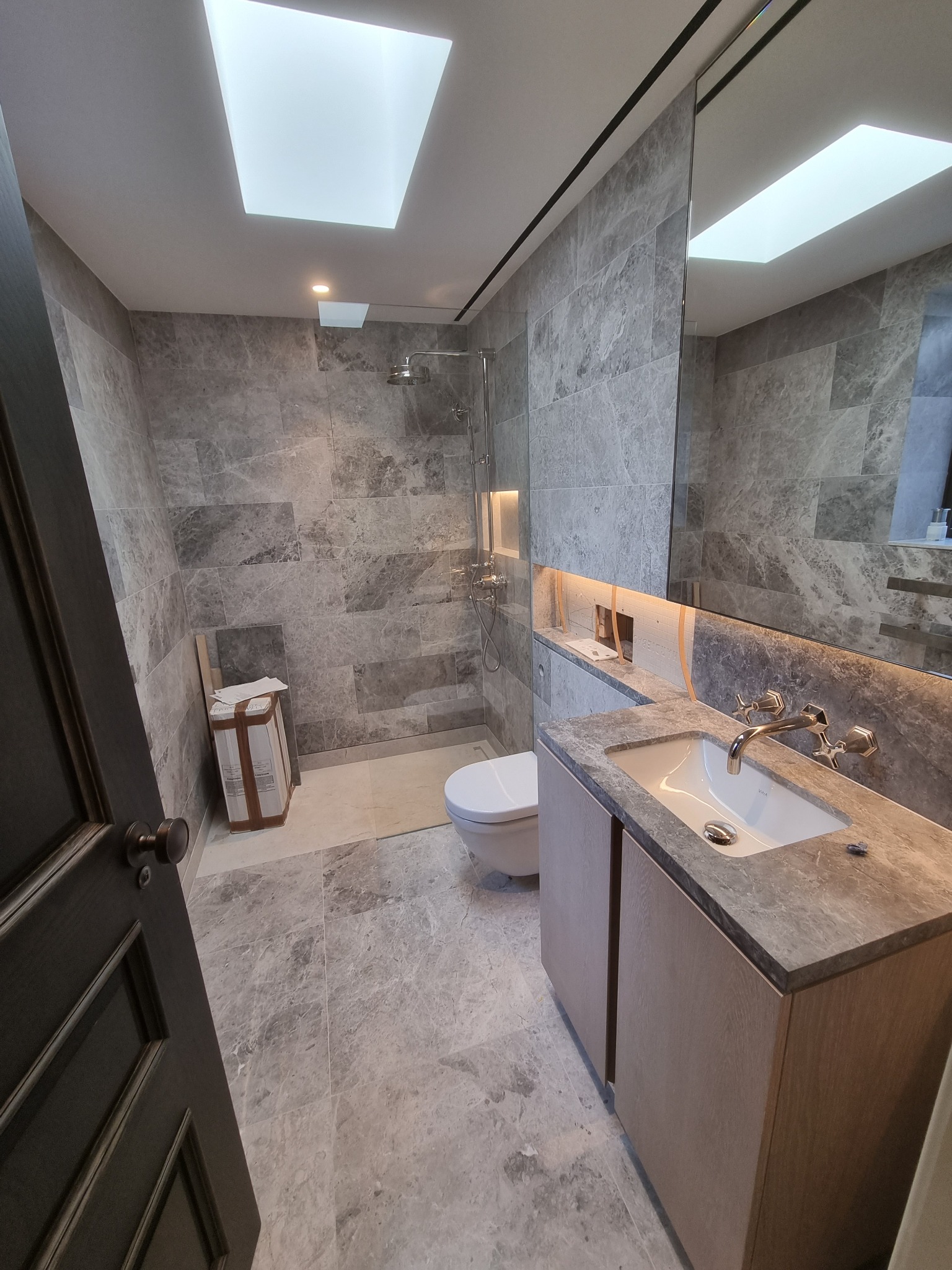 Nowoczesna łazienka z szarymi marmurowymi płytkami, kabiną prysznicową, umywalką z szafką w kolorze drewna i oknem dachowym, częściowo widoczny karton z płytkami.