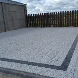 Wykończony podjazd z jasnoszarej kostki brukowej z ciemnoszarym obramowaniem, otoczony betonowym płotem z lewej strony i drewnianym płotem w tle.