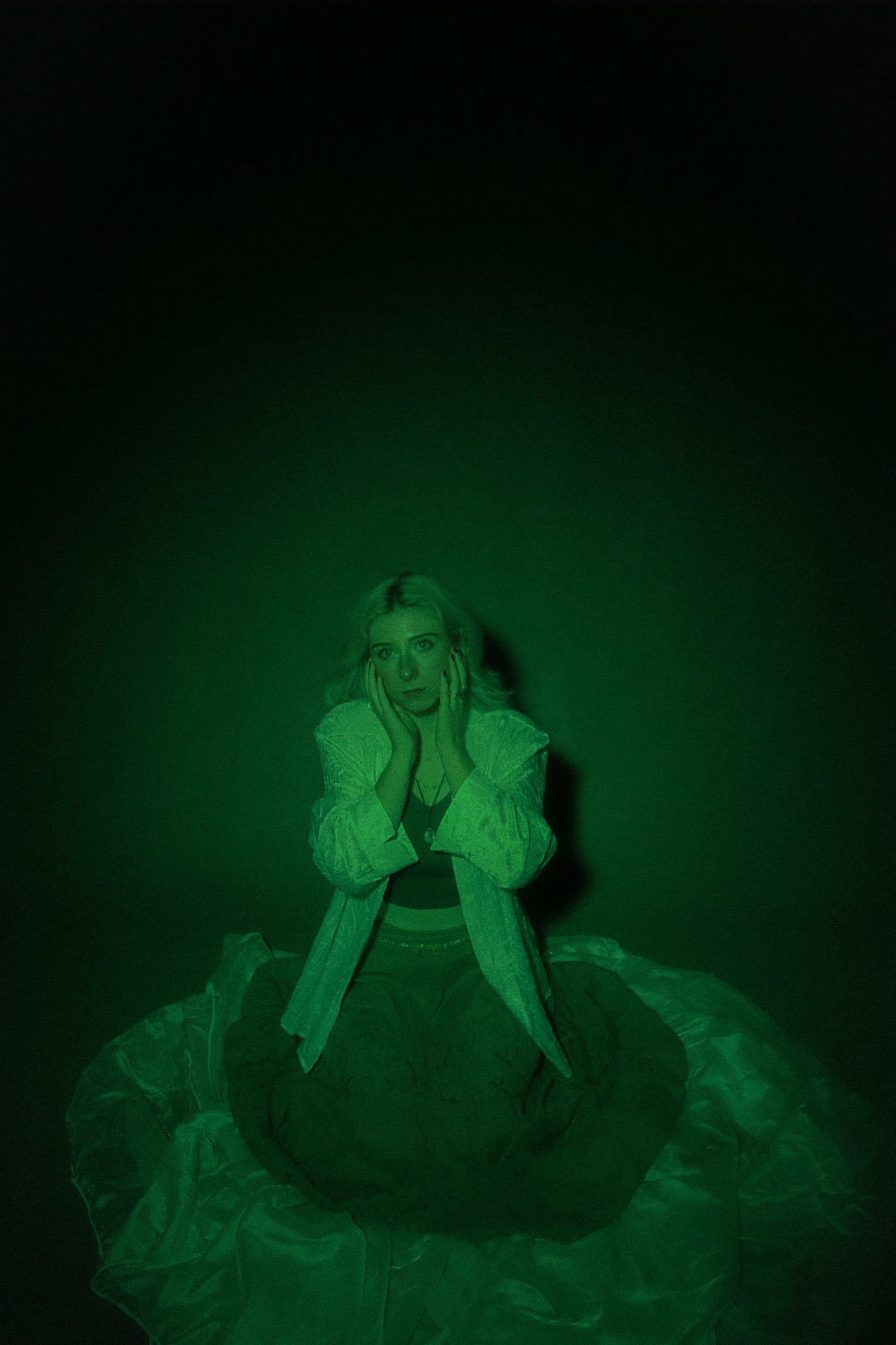 Portret kobiety z blond włosami w zielonym oświetleniu, dłonie przy twarzy, ubrana w bluzkę, spódnicę i luźny płaszcz.