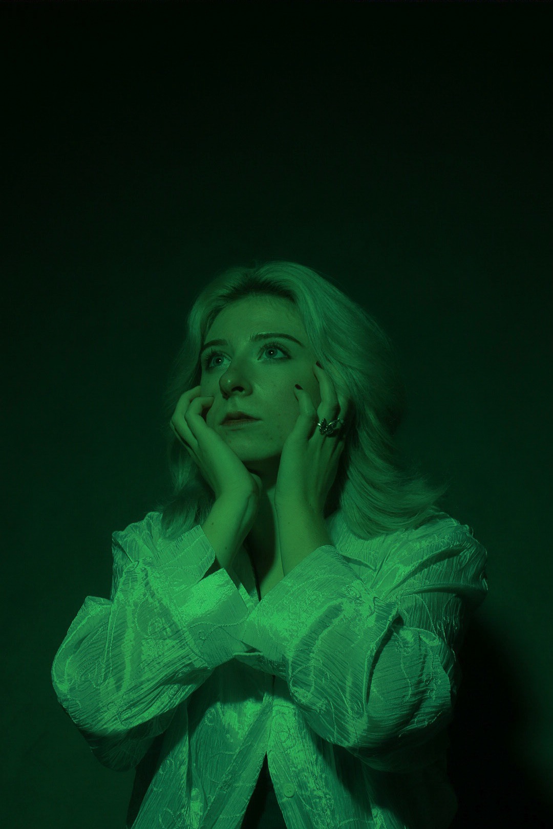 Portret kobiety z blond włosami, zamyślona, opierająca twarz na dłoniach, oświetlona zielonym światłem, widoczny pierścionek na palcu.