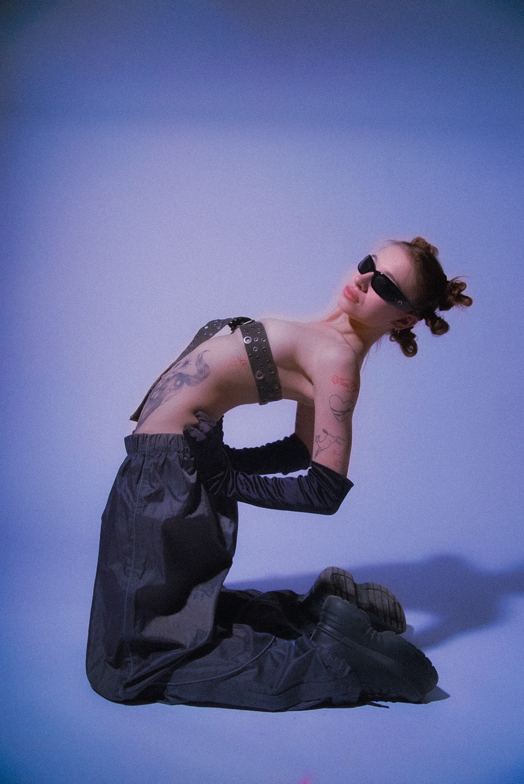 Kobieta w okularach przeciwsłonecznych, czarnych spodniach i rękawiczkach, z tatuażami na ciele, pozuje na kolanach na jednolitym, jasnofioletowym tle.