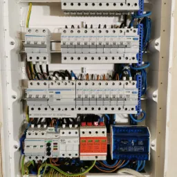 Rozdzielnica elektryczna z zainstalowanymi bezpiecznikami, wyłącznikami różnicowoprądowymi, ogranicznikami przepięć i okablowaniem w białej obudowie, widoczne elementy marki Hager, Tracon...