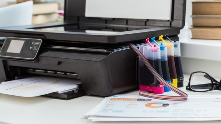 Czarna drukarka atramentowa z zewnętrznym systemem zasilania w tusze, drukująca dokumenty z wykresami, obok okulary i stos książek w tle.