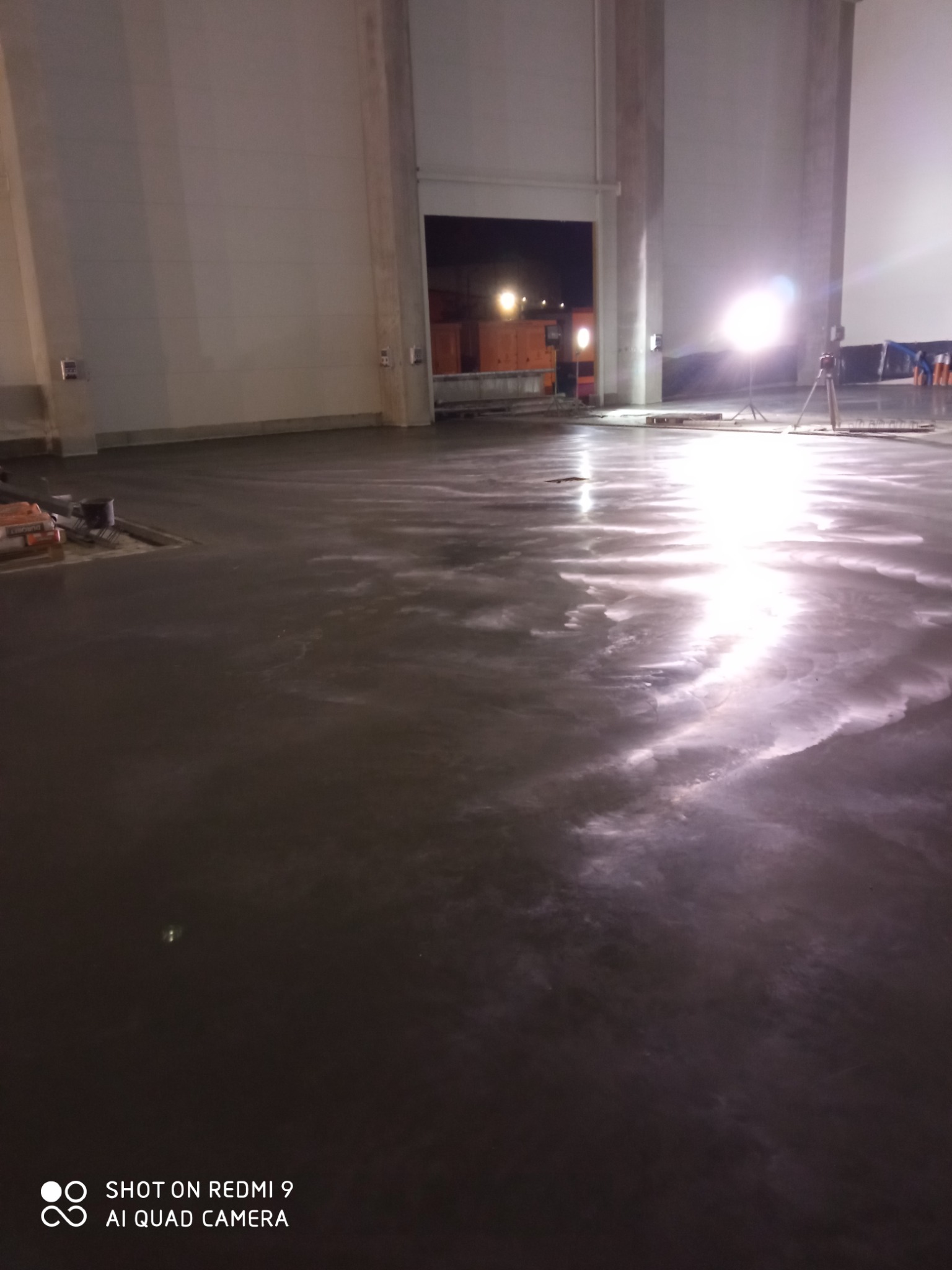 Świeżo wylana, mokra posadzka w hali przemysłowej, widoczne refleksy światła, w tle otwarte wrota i sprzęt budowlany.