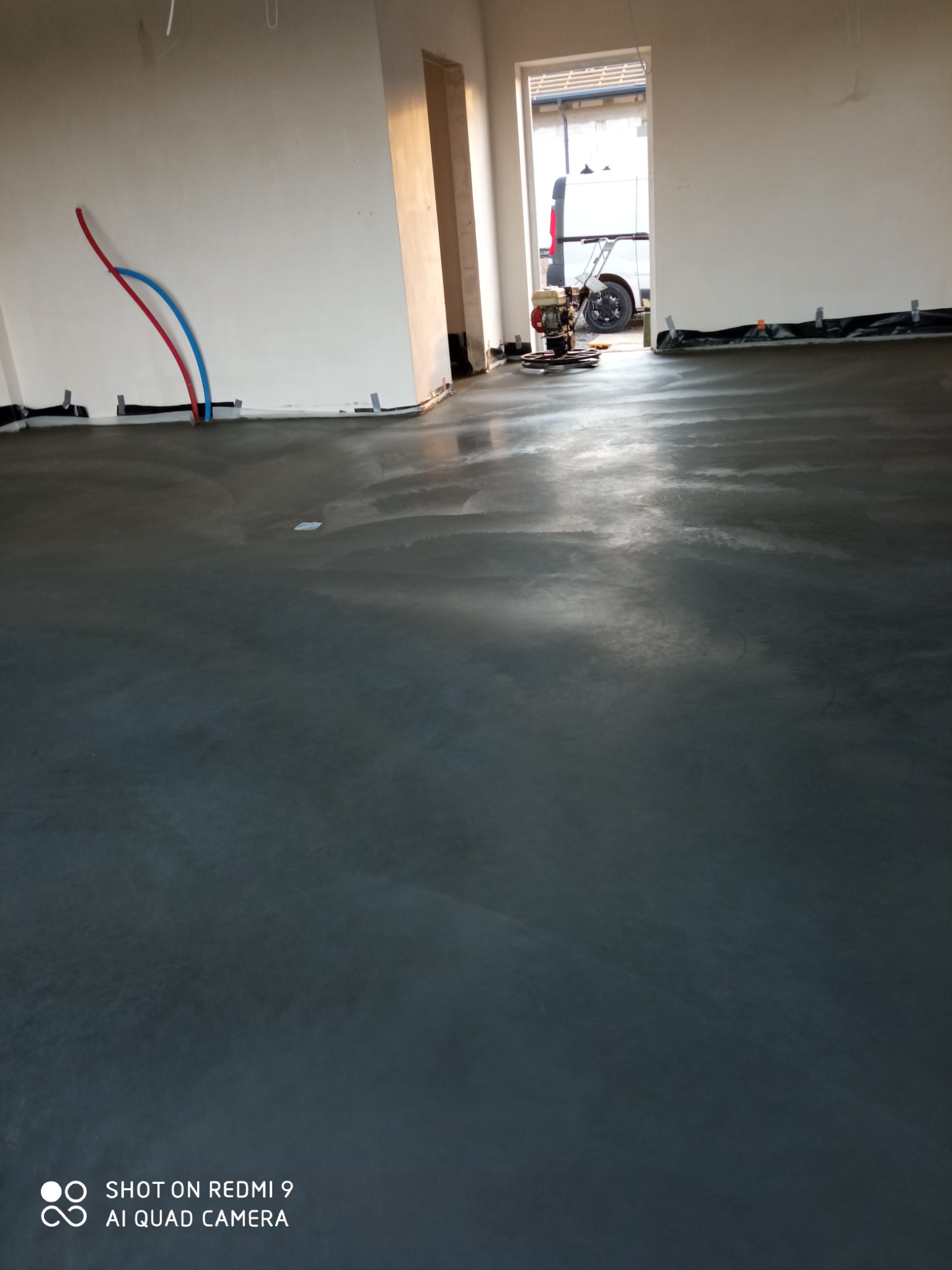 Świeżo wylana, szara posadzka w pomieszczeniu, widoczne wystające rury instalacji grzewczej przy ścianie oraz zacieraczka do betonu w tle, zaparkowana obok białego busa.