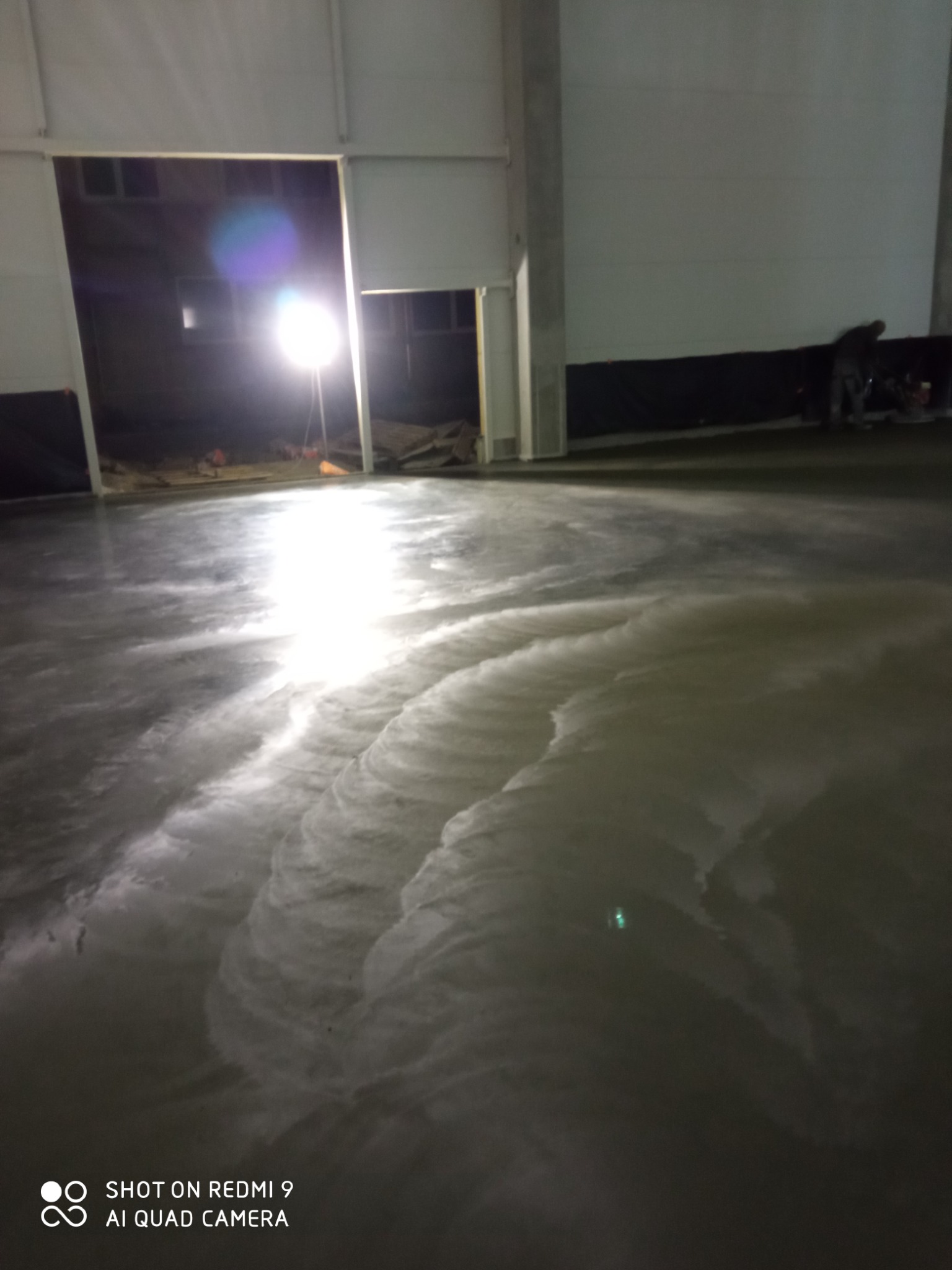 Świeżo wylana, mokra posadzka w hali przemysłowej, oświetlona lampą budowlaną, widoczne ślady po rozprowadzaniu masy.