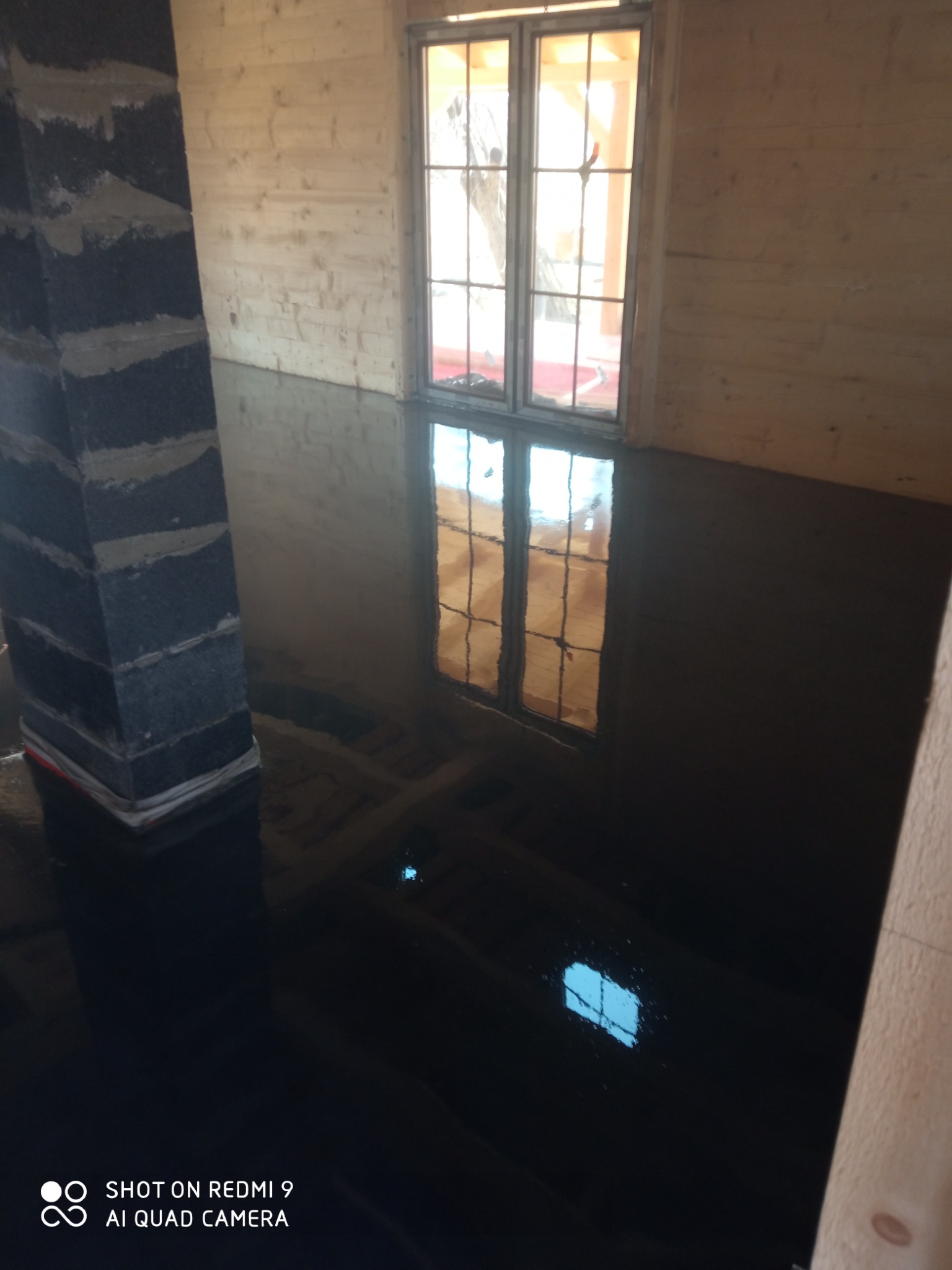 Świeżo wylana, ciemna posadzka w pomieszczeniu z drewnianymi ścianami, odbijająca w oknie widok na zewnątrz, z widocznym filarem w trakcie wykańczania.