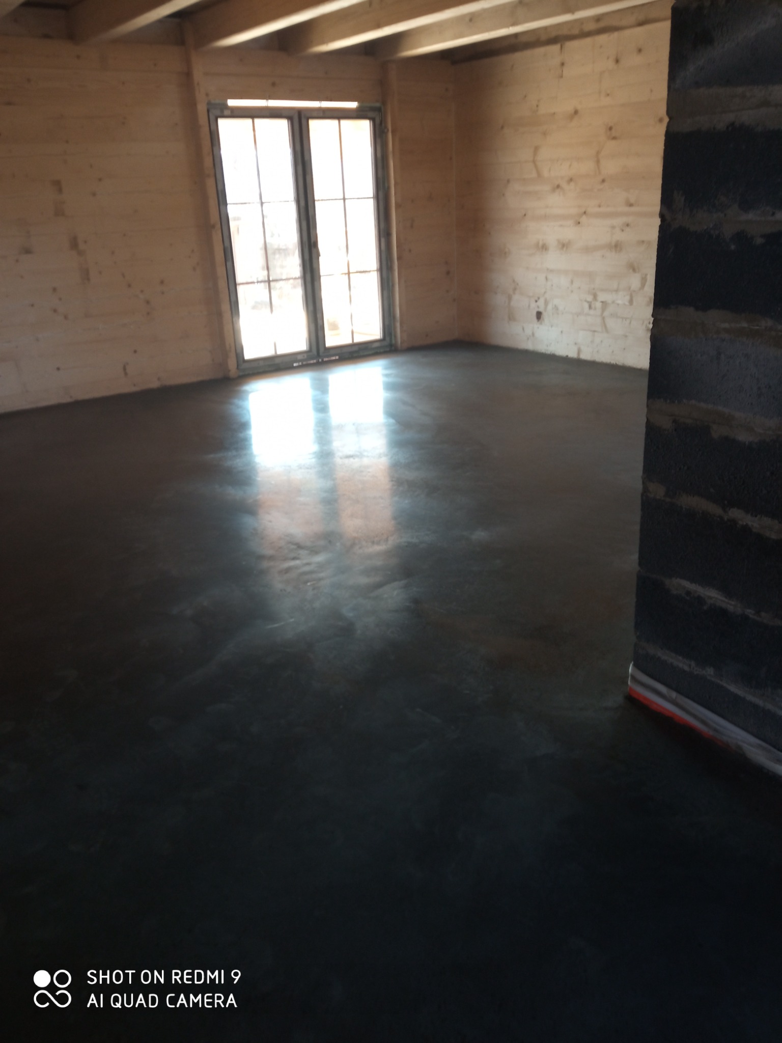 Wnętrze drewnianego domu z wylewką betonową na podłodze, widoczne refleksy światła od okna.