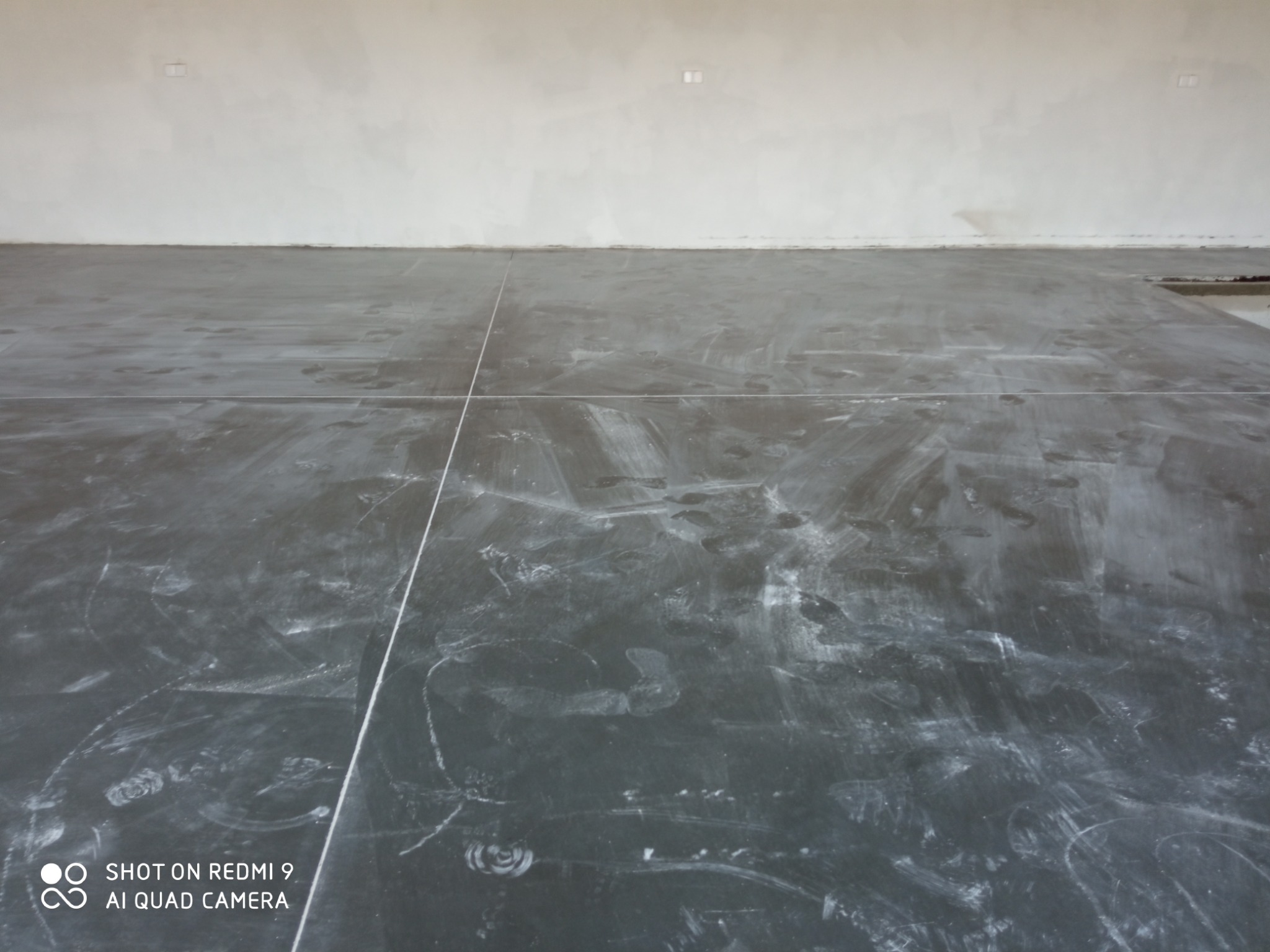 Świeżo wylana, szara posadzka z widocznymi śladami butów i białymi liniami wyznaczającymi podział, surowa ściana w tle.