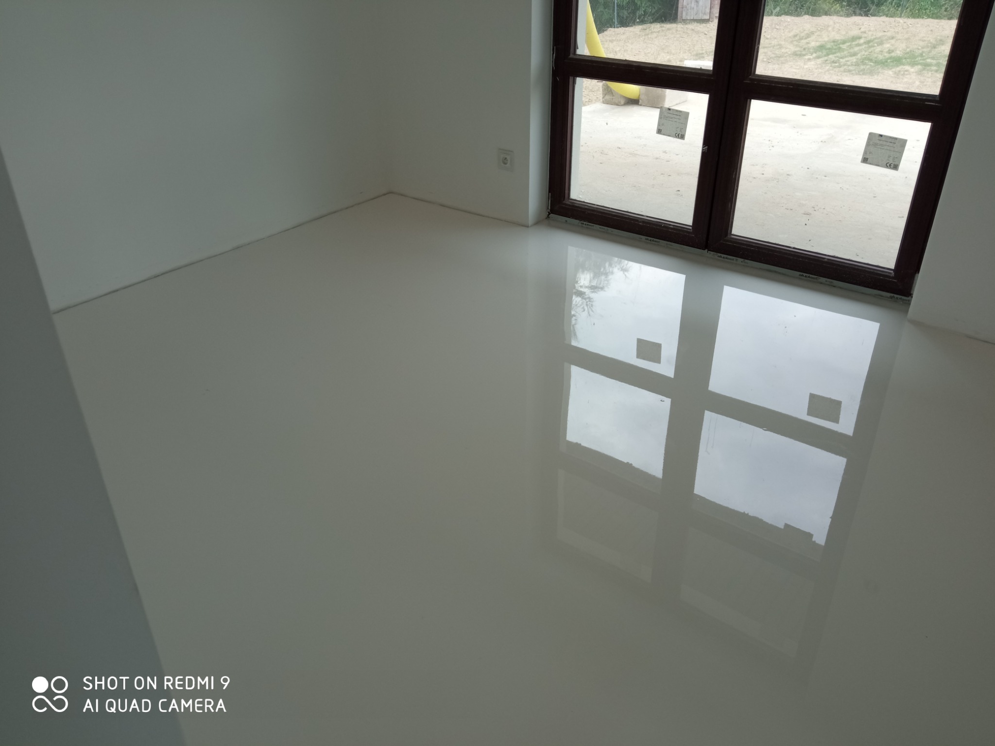 Biała, gładka posadzka w nowym pomieszczeniu z dużym oknem, odbijającym się w powierzchni podłogi, widoczny fragment ściany z gniazdkiem elektrycznym.