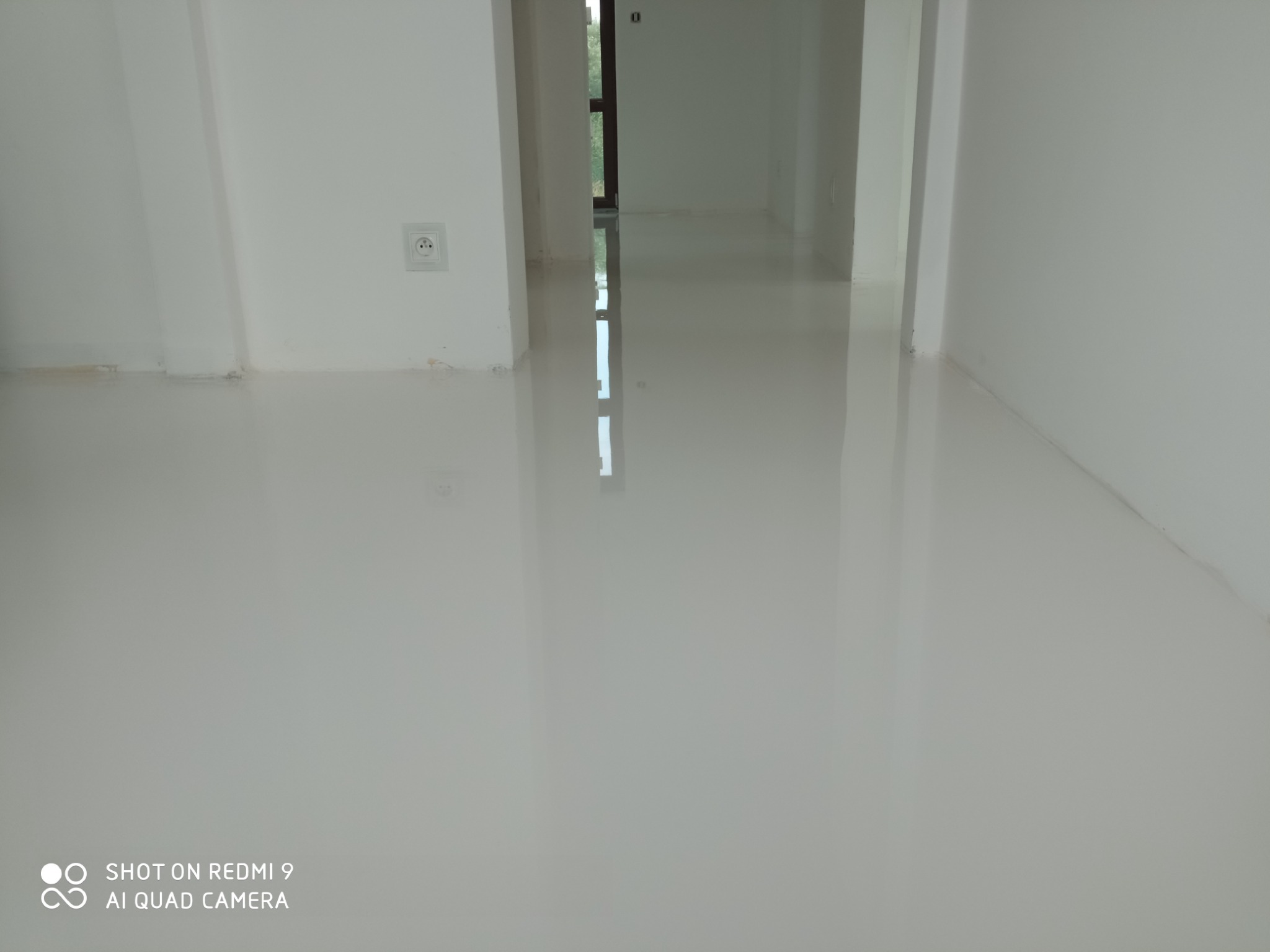 Świeżo wylana, biała, samopoziomująca posadzka w jasnym pomieszczeniu z widocznym gniazdkiem elektrycznym i odbiciem okna w podłodze.