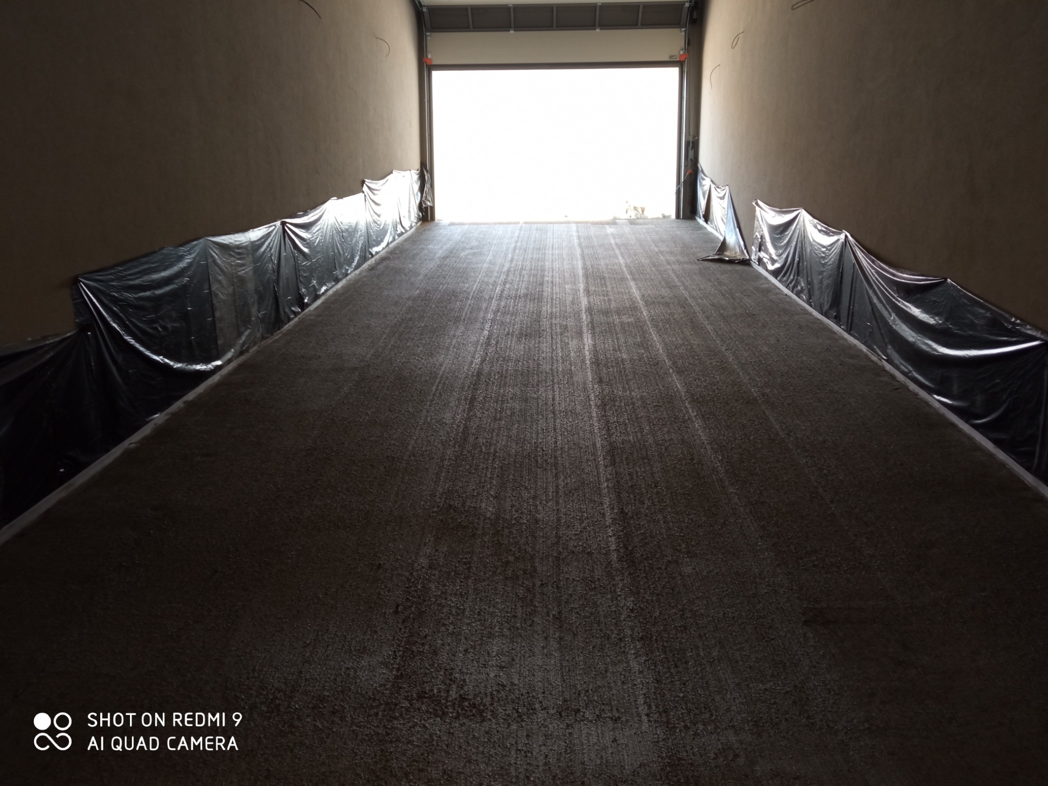 Świeżo wylana, ciemna posadzka jastrychowa w hali produkcyjnej, zabezpieczona folią po bokach, z otwartą bramą wjazdową w tle.