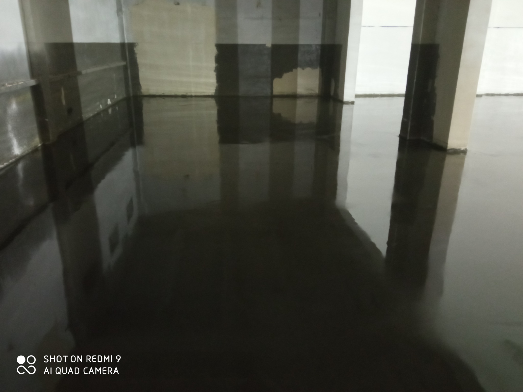 Świeżo wylana, błyszcząca, ciemna posadzka w surowym wnętrzu z widocznymi kolumnami i śladami prac wykończeniowych na ścianach.