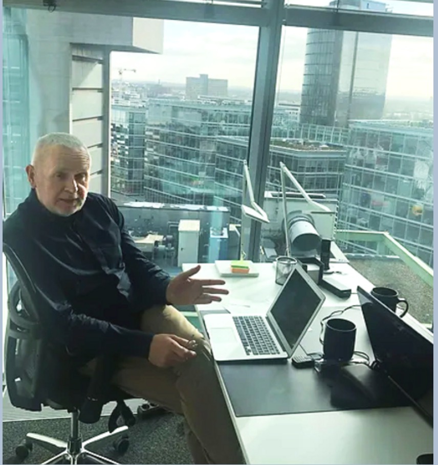 Mężczyzna siedzi na krześle biurowym w nowoczesnym biurze z widokiem na miasto, na biurku laptop, lampka i kubki.