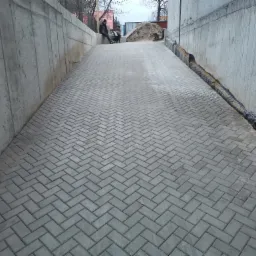 Ukończona ścieżka z szarej kostki brukowej ułożonej w jodełkę, ograniczona betonowymi ścianami i siatką drenażową, z dwoma osobami siedzącymi w oddali.