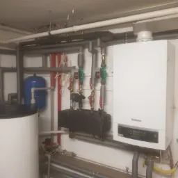 Biały kondensacyjny piec gazowy marki Buderus zamontowany na ścianie, z widocznymi rurami miedzianymi i szarymi, oraz zasobnikiem ciepłej wody i niebieskim naczyniem przeponowym w tle.