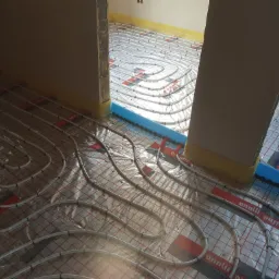 Wnętrze w trakcie montażu ogrzewania podłogowego: białe rurki grzewcze ułożone w pętle na folii z siatką, przymocowane klipsami, z widoczną niebieską izolacją brzegową przy ścianie.