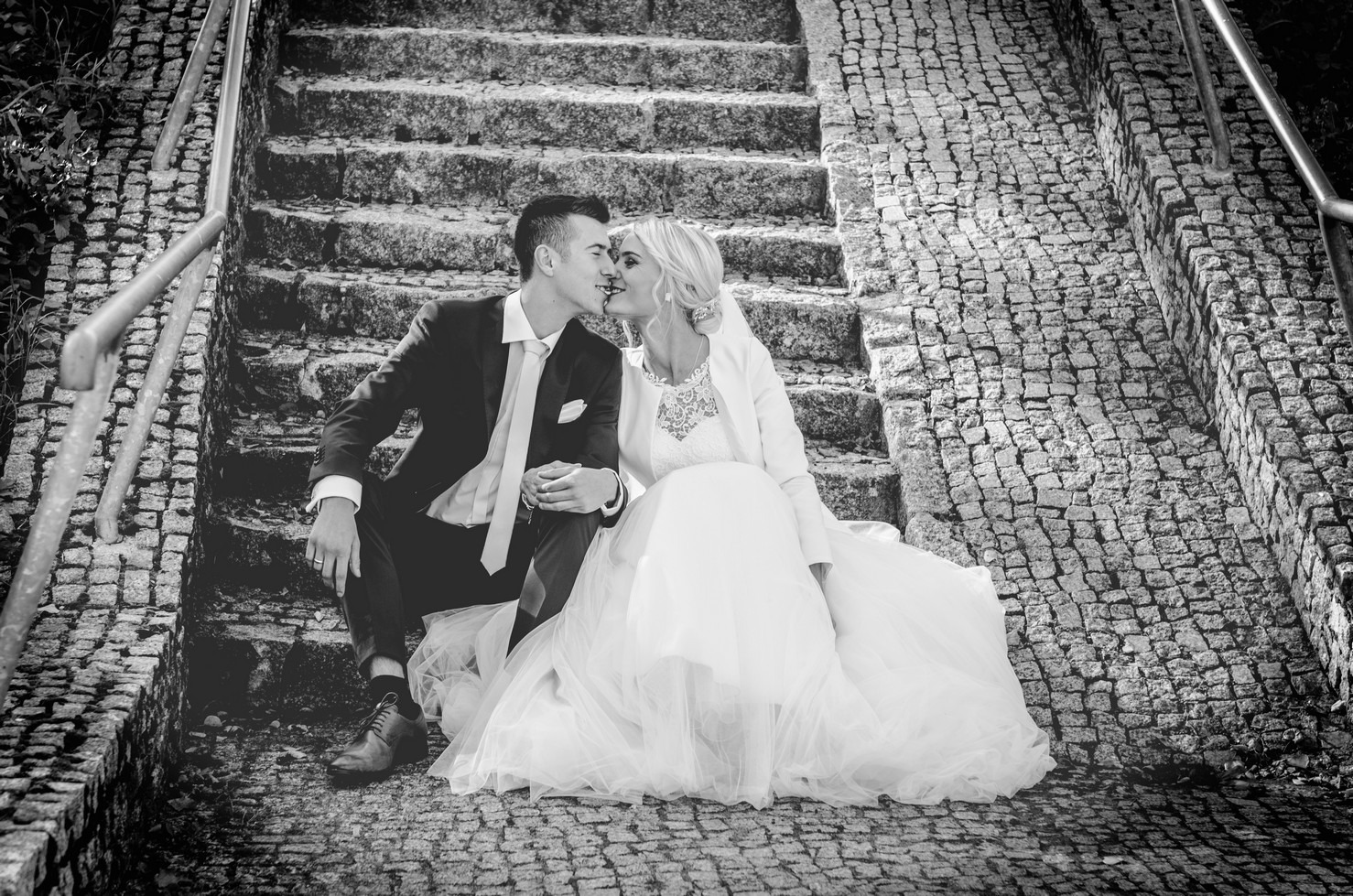 Czarno-białe zdjęcie całującej się pary młodej siedzącej na kamiennych schodach z metalową barierką, panna młoda w długiej sukni i bolerku, pan młody w ciemnym garniturze i jasnym krawacie.