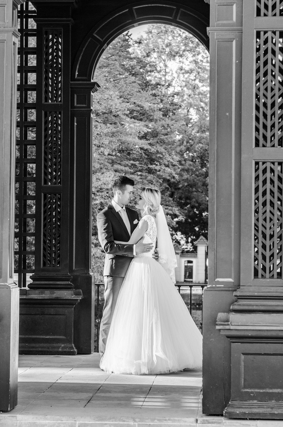 Czarno-białe zdjęcie narzeczonej w koronkowej sukni i welonie, obejmowanej przez pana młodego w garniturze, na tle ażurowej konstrukcji altany.