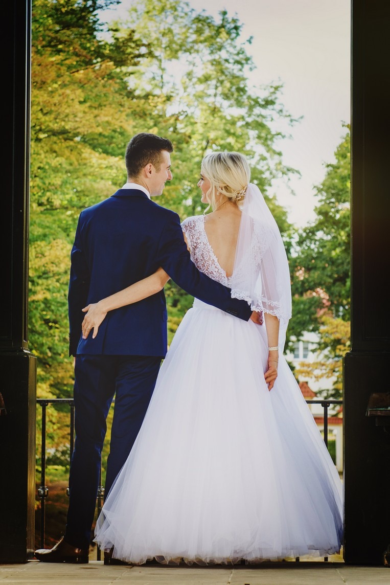 Para młoda, widok z tyłu, w objęciach na tle zieleni, panna młoda w sukni z koronkowymi plecami i welonem, pan młody w granatowym garniturze.