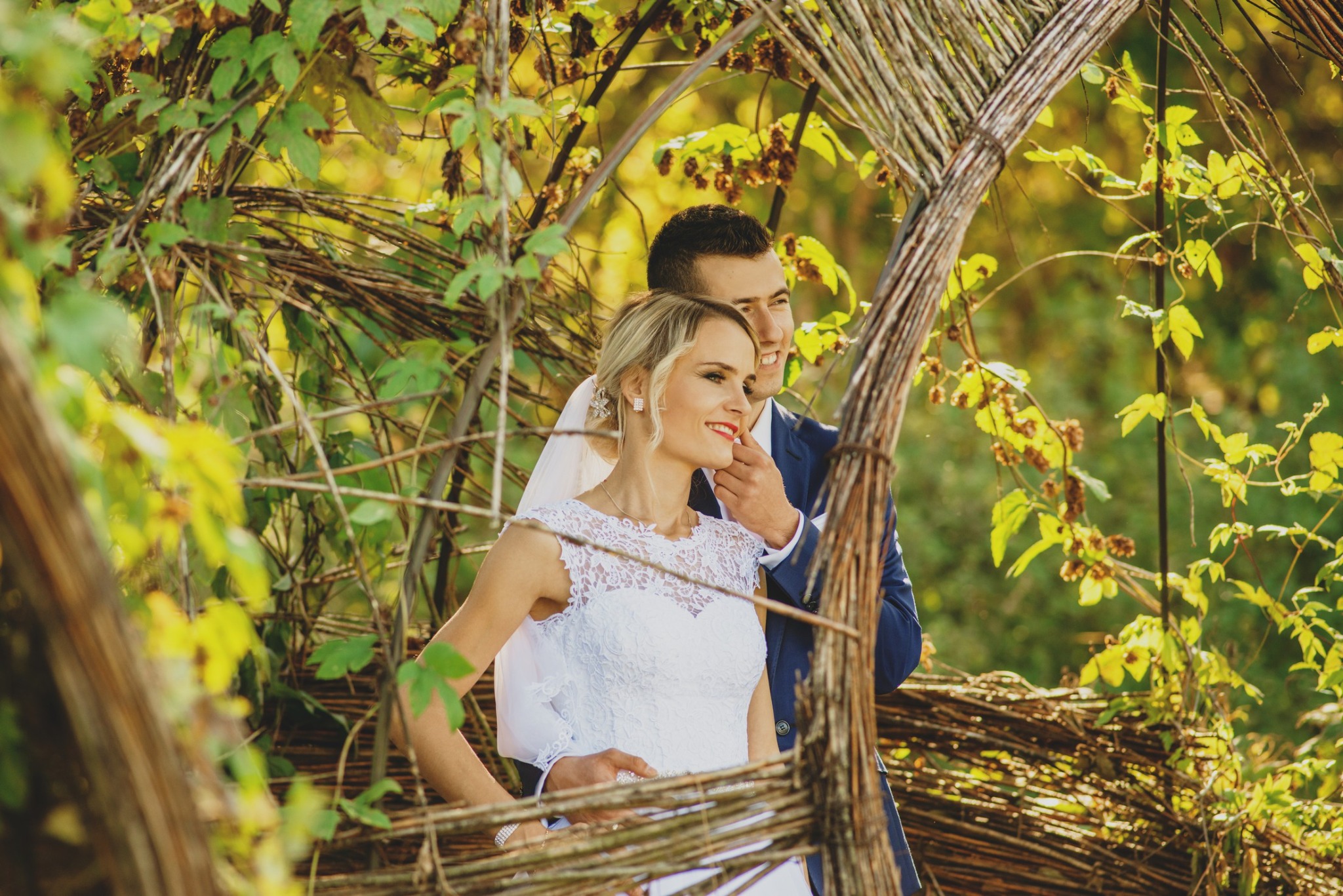 Uśmiechnięta para młoda w strojach ślubnych, pozująca w plenerze, otoczona gałęziami i liśćmi w słoneczny dzień.
