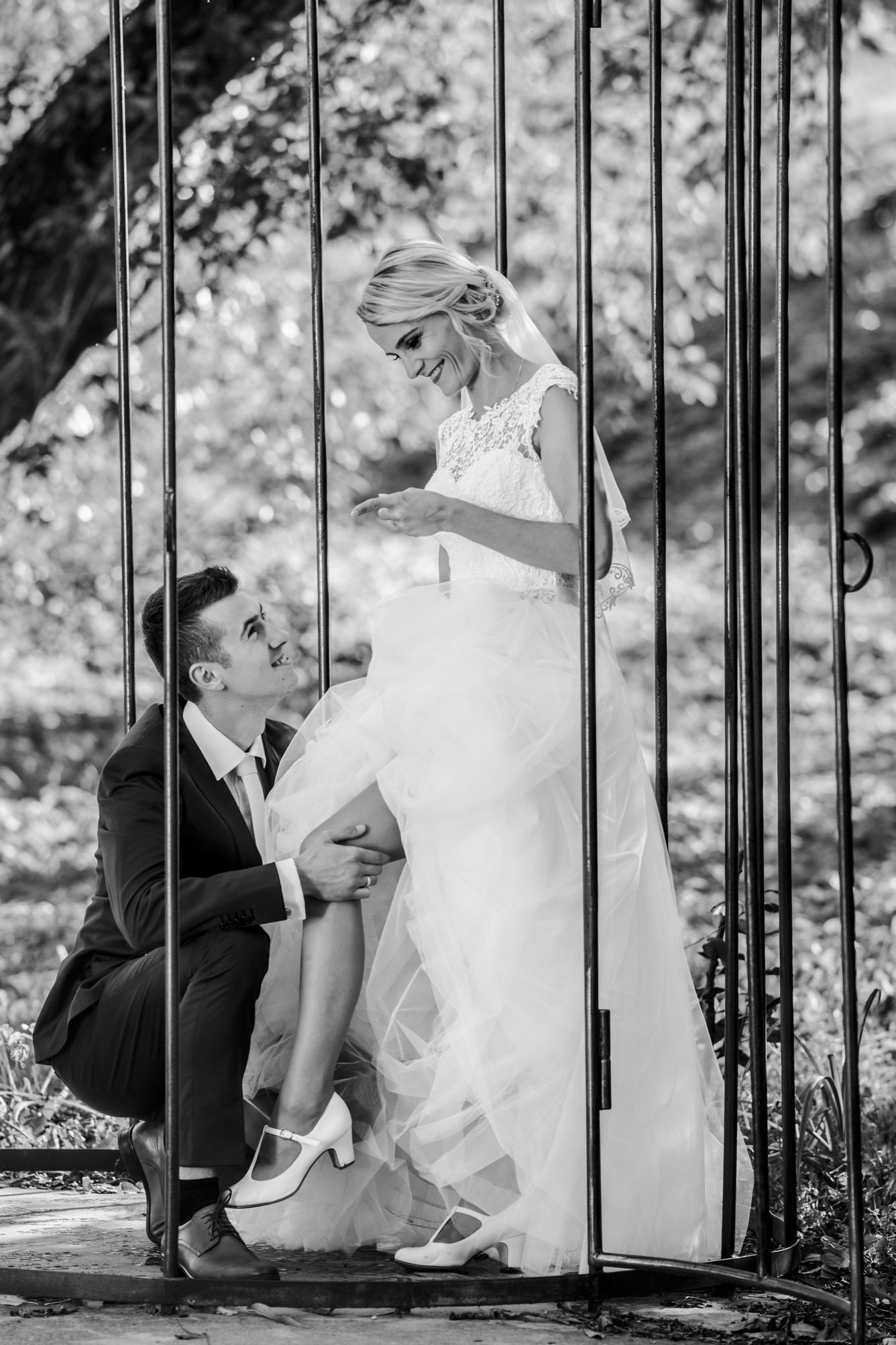 Czarno-białe zdjęcie narzeczonej w koronkowej sukni i butach w stylu retro, siedzącej na kolanach narzeczonego w garniturze, oboje uśmiechnięci, za metalową konstrukcją przypominającą klatkę...