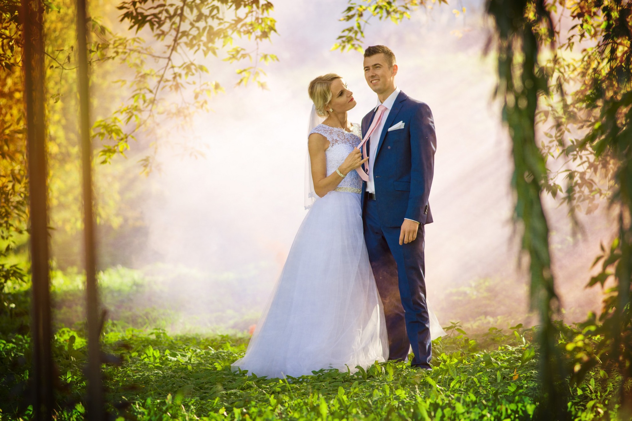 Para młoda w lesie: Panna młoda w koronkowej sukni i welonie delikatnie poprawia krawat pana młodego w granatowym garniturze na tle mglistego, leśnego krajobrazu.