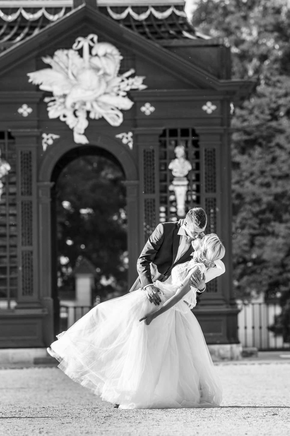 Czarno-białe zdjęcie całującej się pary młodej, panna młoda w sukni balowej, pan młody w garniturze, na tle ozdobnej altany z rzeźbami.