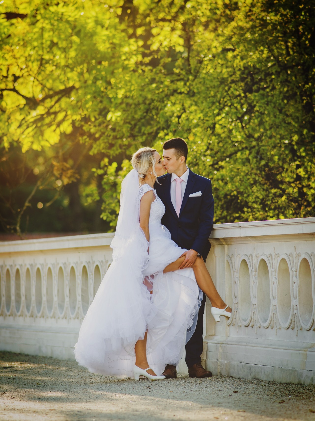 Pocałunek młodej pary w plenerze: panna młoda w białej sukni i welonie, oparta o kamienną balustradę, pan młody w granatowym garniturze i różowym krawacie, zielone drzewa w tle, ciepłe światło...