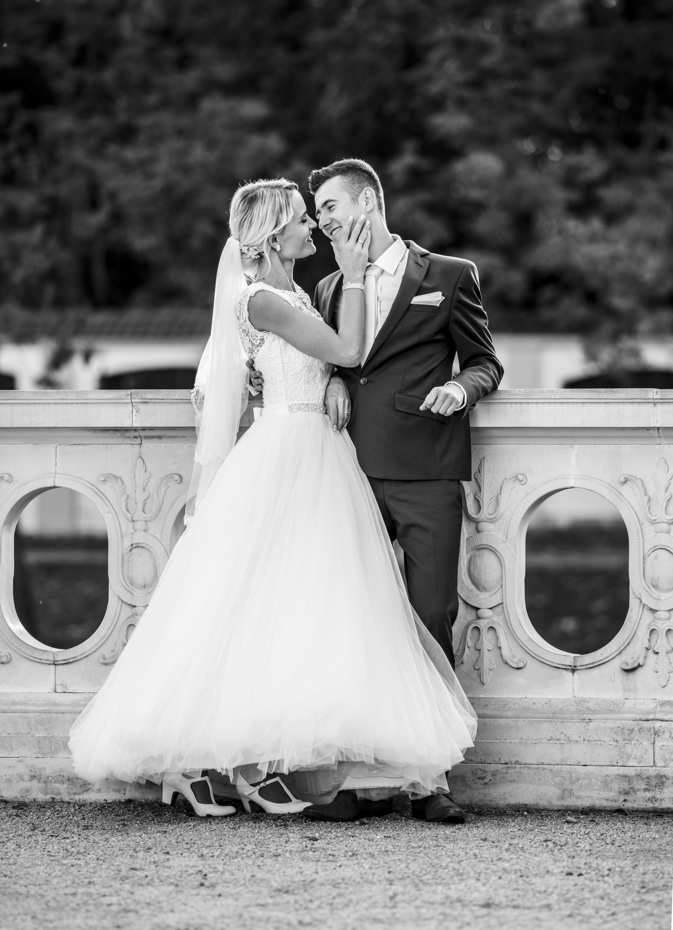 Czarno-białe zdjęcie pary młodej: panna młoda w koronkowej sukni i welonie dotyka twarzy pana młodego w garniturze, oboje uśmiechają się do siebie, opierając się o ozdobną balustradę.