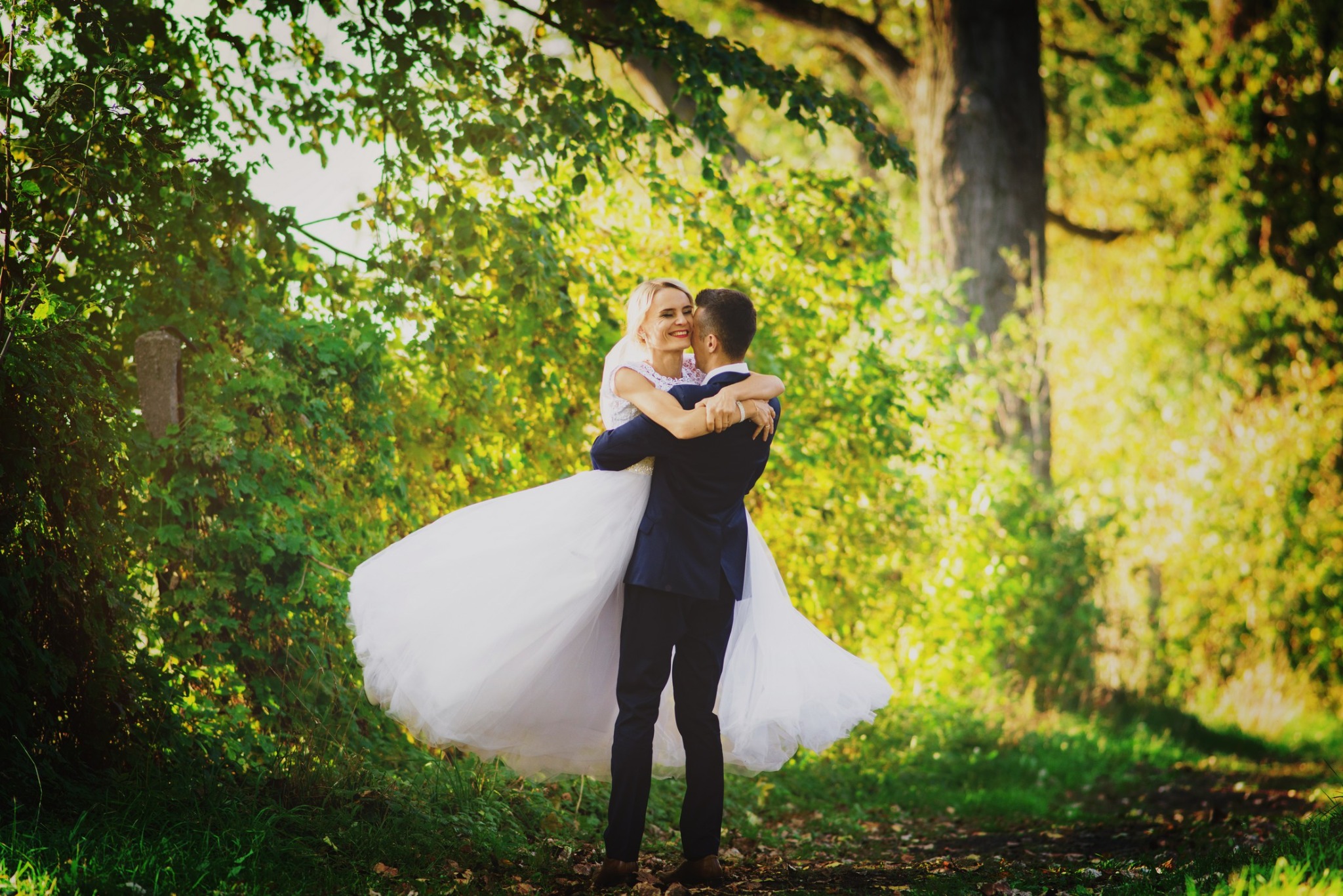 Uśmiechnięta panna młoda w białej sukni, unoszona przez pana młodego w granatowym garniturze na tle zieleni drzew i ścieżki leśnej w słoneczny dzień.