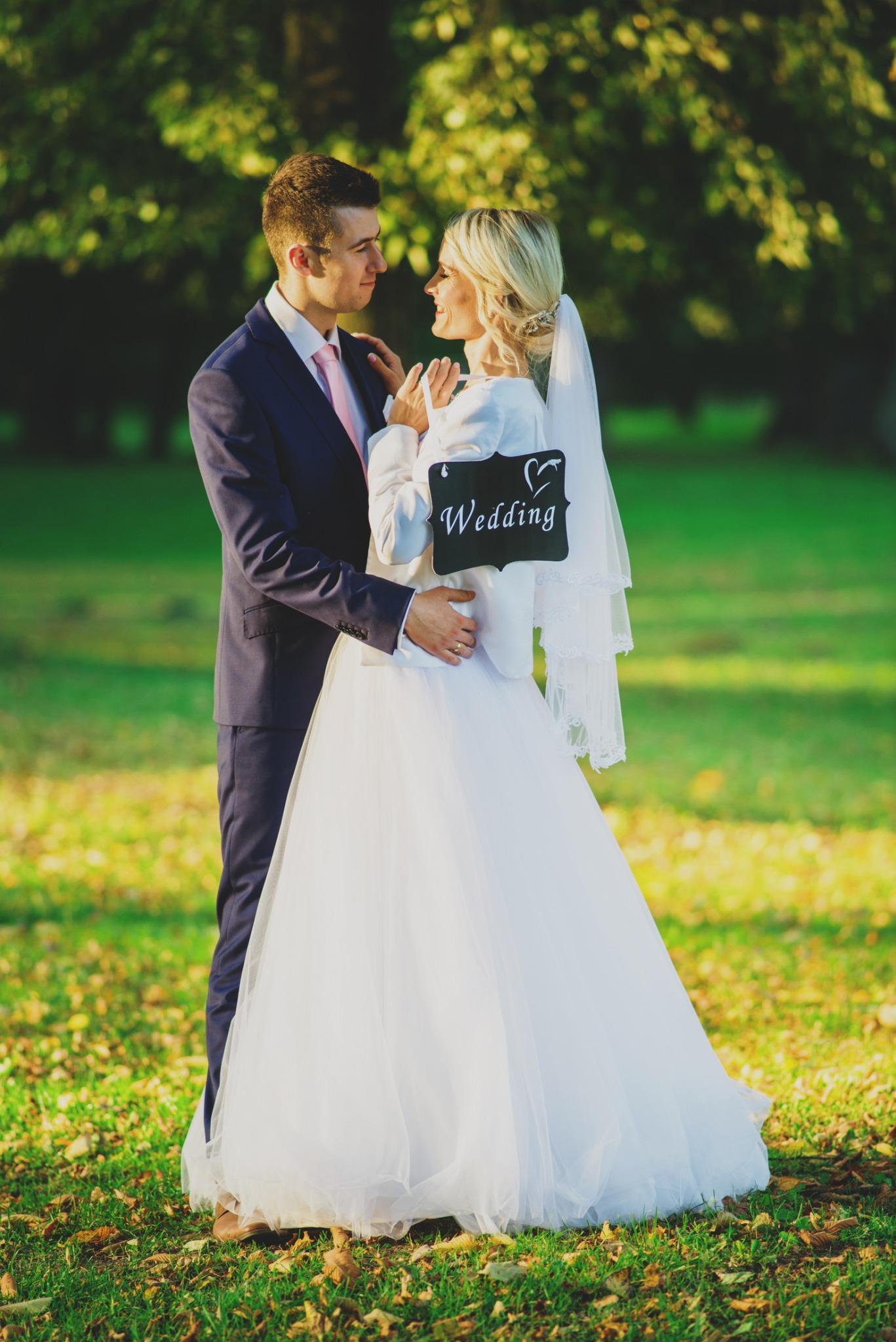 Para młoda w plenerze, panna młoda trzyma tabliczkę z napisem 'Wedding' i sercem, delikatne światło słoneczne.