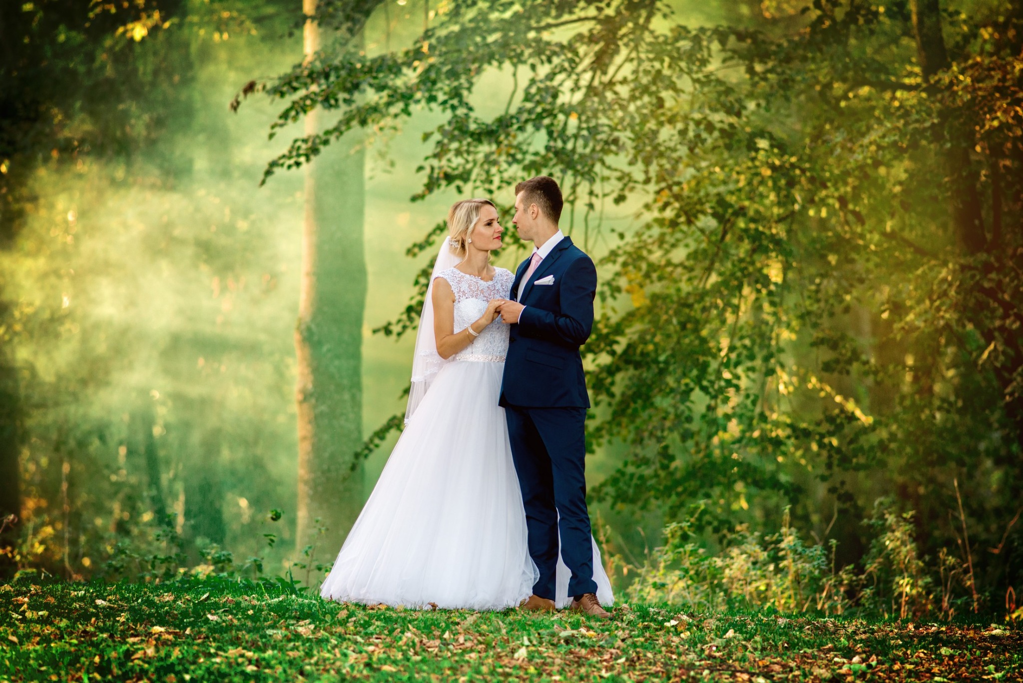 Para młoda w lesie, panna młoda w białej sukni z koronkową górą i welonem, pan młody w granatowym garniturze, trzymają się za ręce, delikatne światło słoneczne przesącza się przez drzewa.