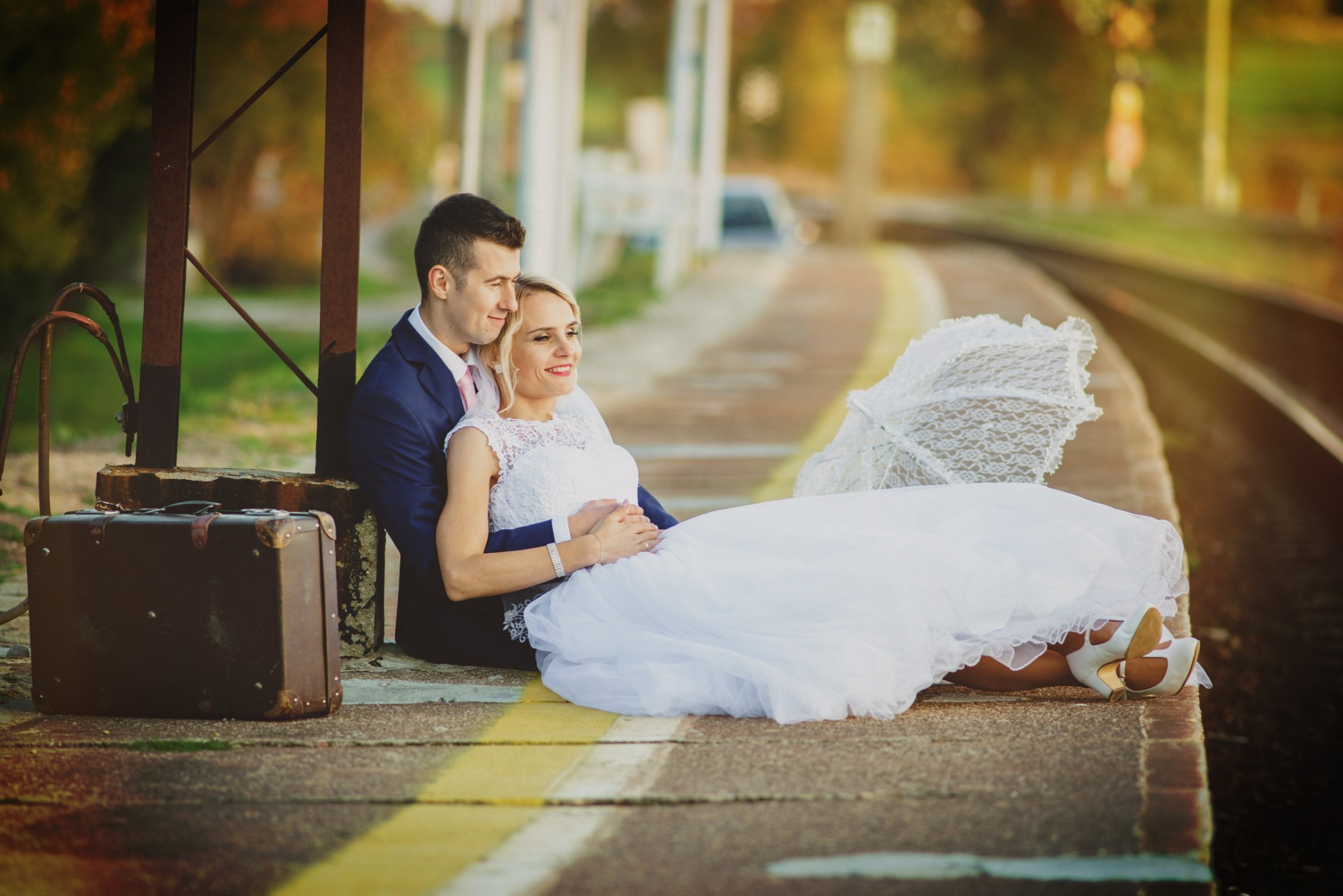 Para młoda siedzi na peronie kolejowym, obok stoi walizka, panna młoda trzyma koronkowy parasol