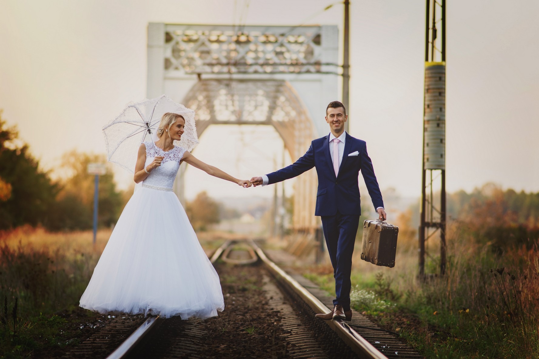 Para młoda trzymająca się za ręce, idąca po torach kolejowych; panna młoda w białej sukni z koronkową górą i parasolką, pan młody w granatowym garniturze z walizką; w tle most kolejowy i jesienny...