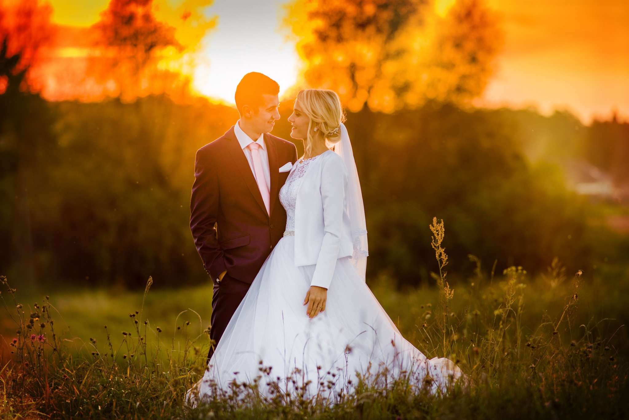 Para młoda w sukni ślubnej i garniturze, patrząca na siebie na tle zachodzącego słońca, w plenerze z wysoką trawą.