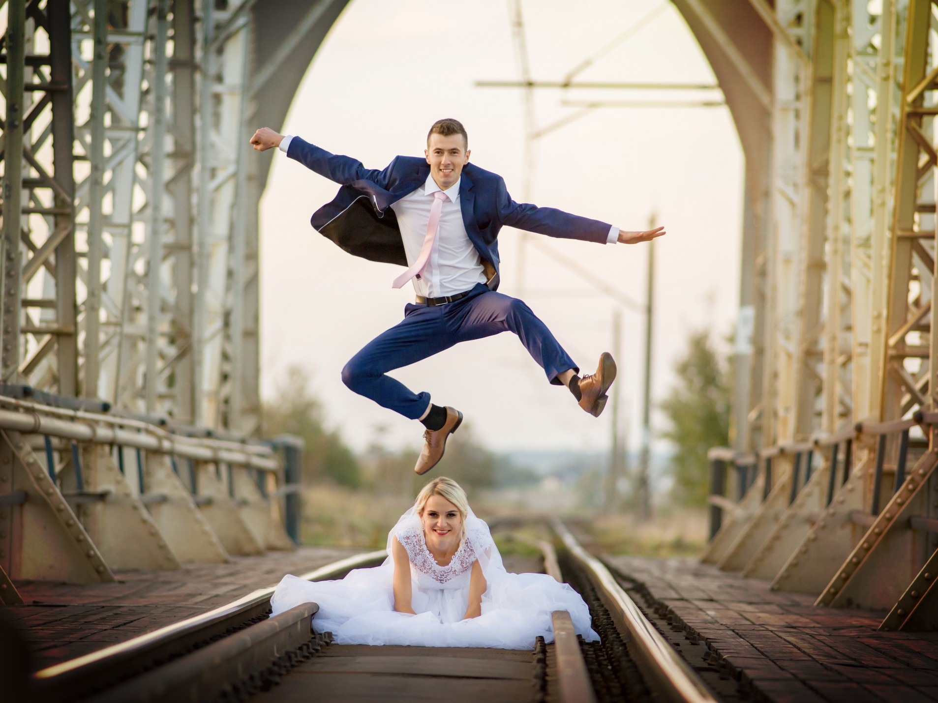 Para młoda na torach kolejowych: panna młoda klęczy w białej sukni, pan młody skacze w garniturze, z uśmiechem na twarzy.