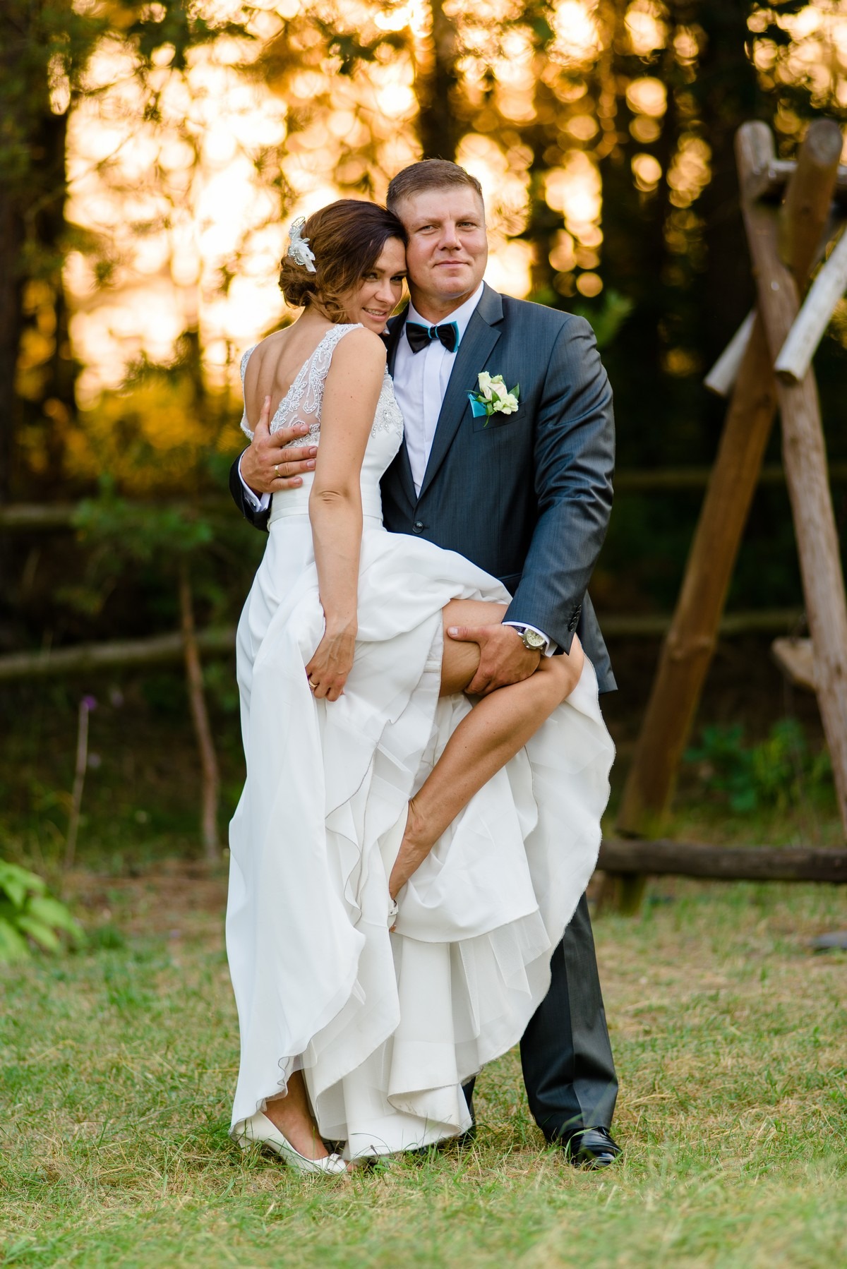 Uśmiechnięta para młoda: panna młoda w białej sukni z koronkową górą i upiętymi włosami z kwiatem, trzymana przez pana młodego w szarym garniturze z muchą i butonierką, na tle drzew i drewnianej...