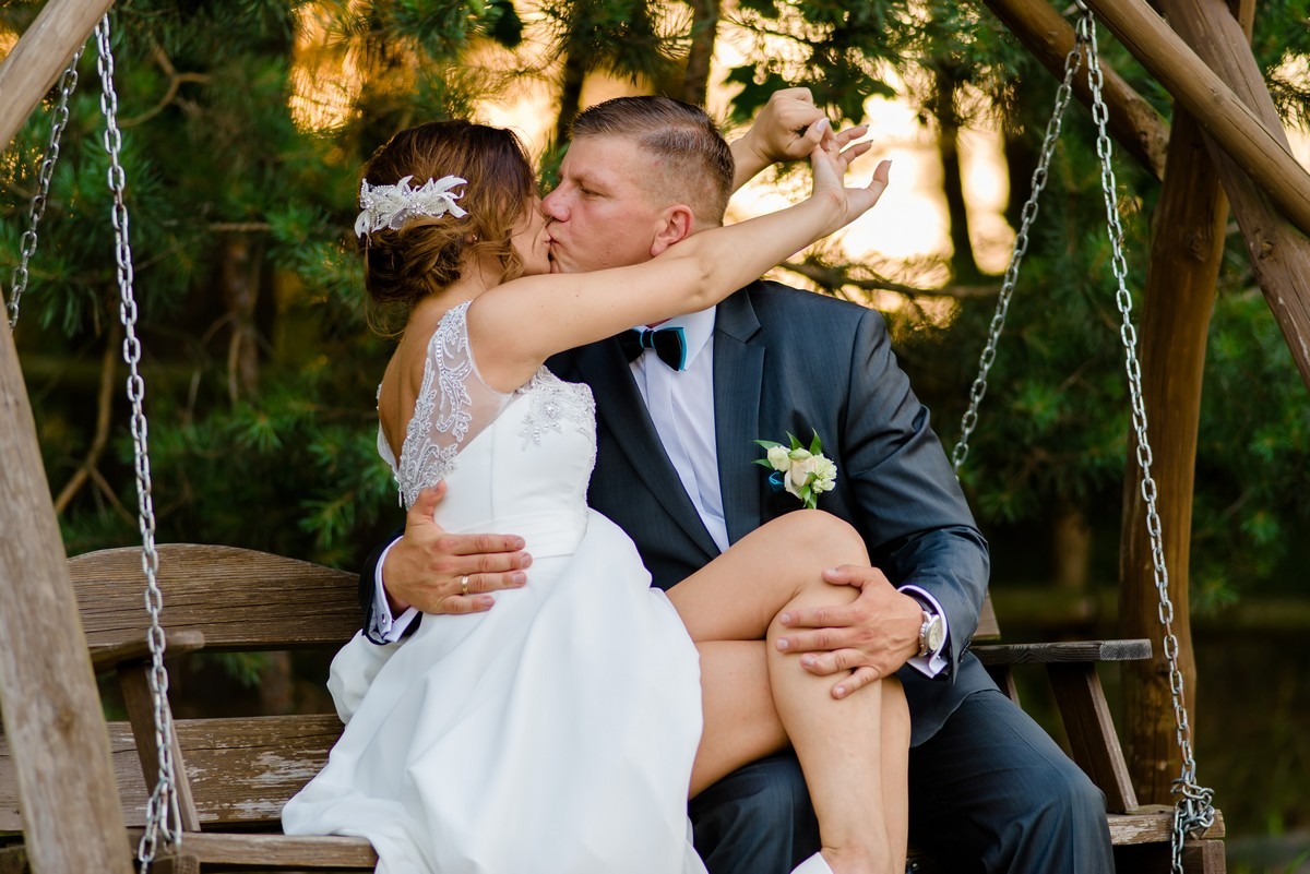 Pocałunek pary młodej siedzącej na drewnianej huśtawce w plenerze, ona w białej sukni z koronkowymi zdobieniami, on w szarym garniturze z muchą i butonierką.