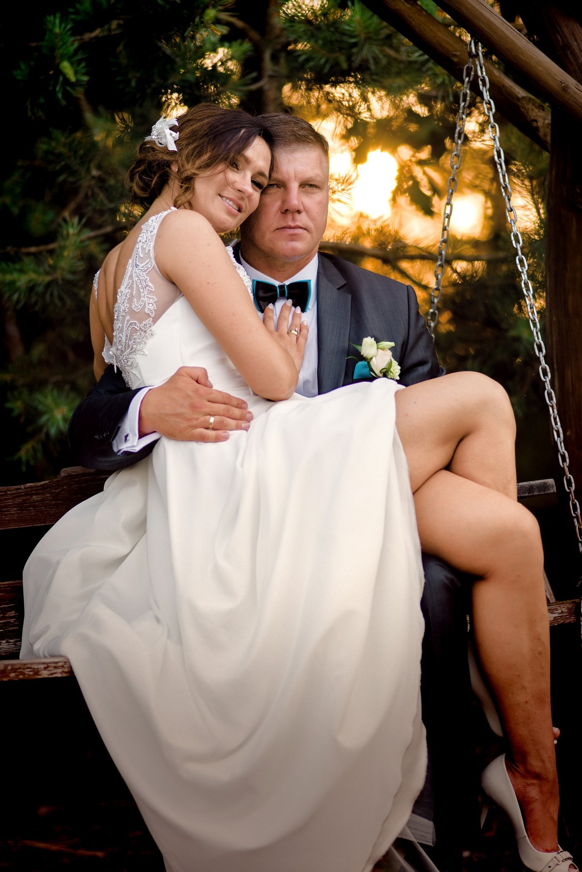 Para młoda siedząca na drewnianej huśtawce: panna młoda w białej sukni z koronkowymi plecami i pan młody w szarym garniturze z turkusową muchą, w tle zieleń i zachodzące słońce.