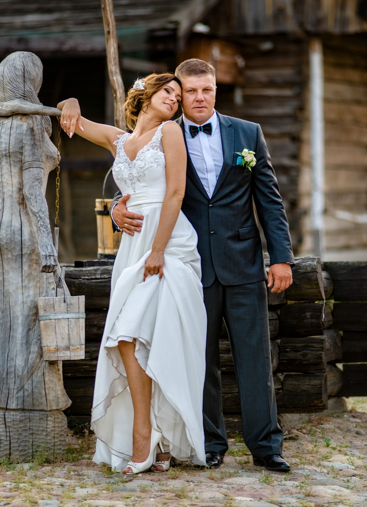 Para młoda w strojach ślubnych pozuje przy drewnianej studni z rzeźbą kobiety, panna młoda opiera się o rzeźbę, a pan młody obejmuje ją w talii.