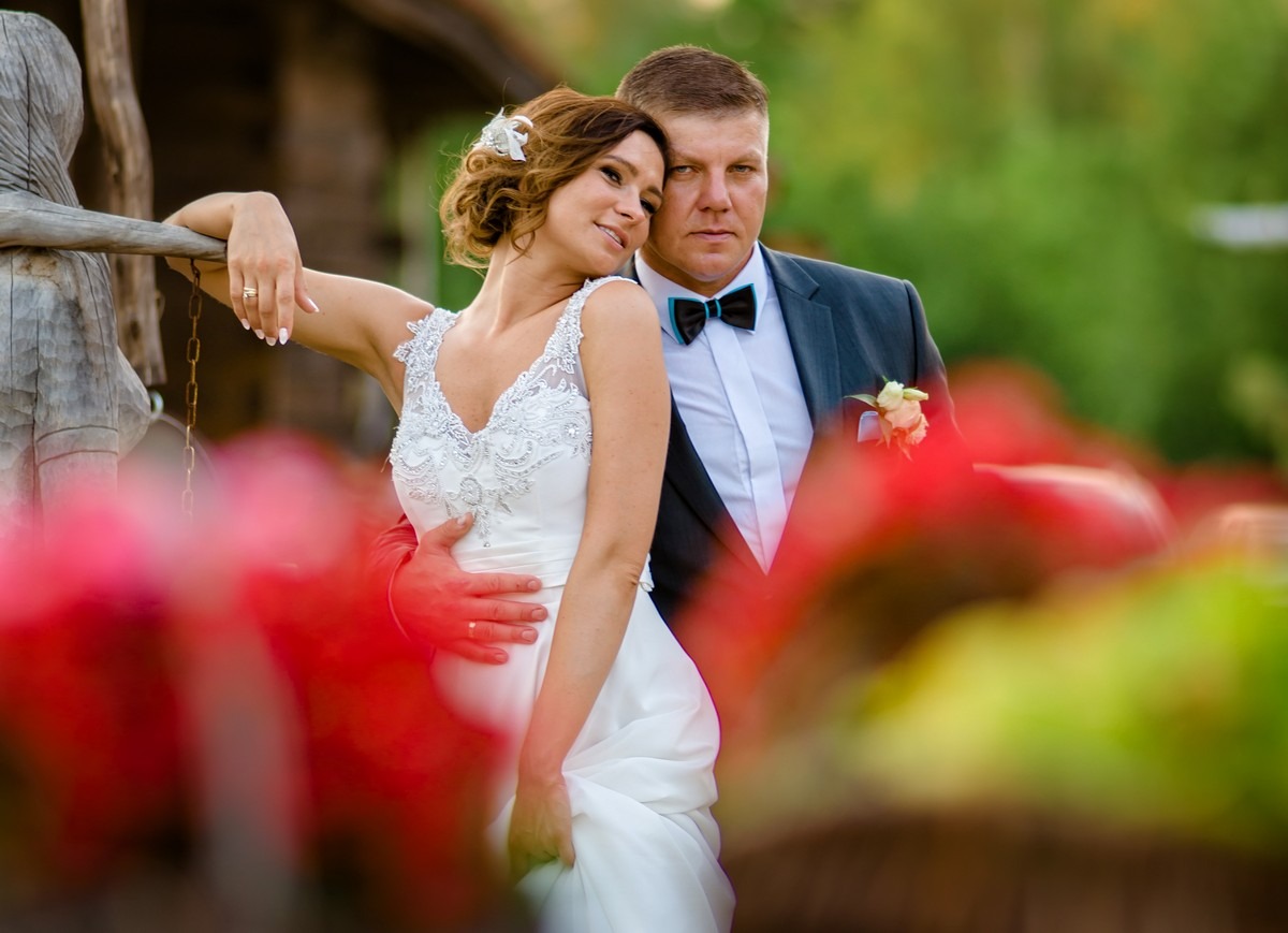 Para młoda w objęciach, panna młoda w białej sukni z koronkowymi zdobieniami i kwiatem we włosach, pan młody w ciemnym garniturze z turkusową muchą, pozują na tle zieleni i czerwonych kwiatów...