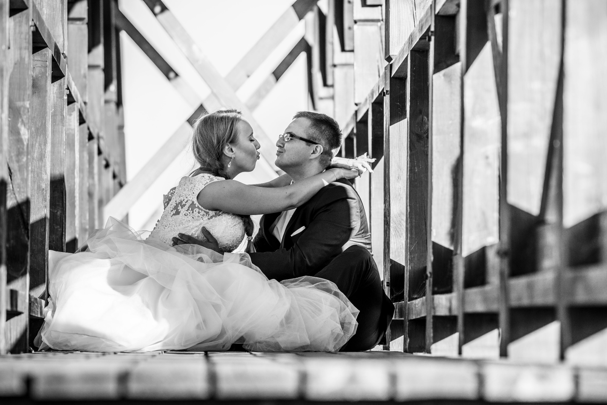 Czarno-białe zdjęcie pary młodej siedzącej na drewnianym moście, panna młoda w koronkowej sukni i welonie, pan młody w garniturze i okularach, w uścisku, z twarzami blisko siebie.