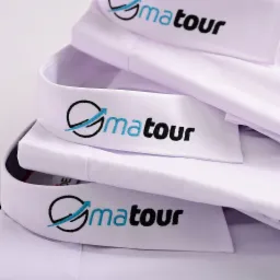 Stos białych koszul z wyhaftowanym logo 'Gmatour' w kolorze czarnym i błękitnym, widoczne kołnierzyki.