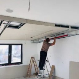 Finma remontowo budowlana - Pracownik na drabinie montuje konstrukcję sufitu podwieszanego z płyt kartonowo-gipsowych w pomieszczeniu z oknem, widoczne profile metalowe i częściowo zamontowane płyty.