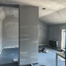 Wnętrze w trakcie remontu: ściany i sufit pokryte świeżo nałożoną gładzią, widoczne puszki farby Dekoral i narzędzia malarskie na podłodze zabezpieczonej folią.