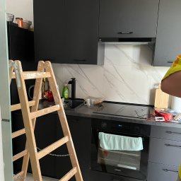 Finma remontowo budowlana - Nowoczesna, czarna zabudowa kuchenna z marmurowym backsplashiem, drabina malarska oparta o szafki, sugerująca prace wykończeniowe.