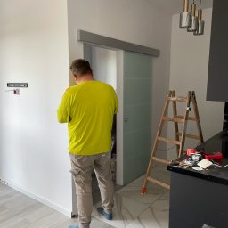 Finma remontowo budowlana - Pracownik w jaskrawej koszulce montuje listwę przypodłogową przy szklanych drzwiach przesuwnych w wyremontowanym wnętrzu, widoczna drabina i narzędzia na blacie.