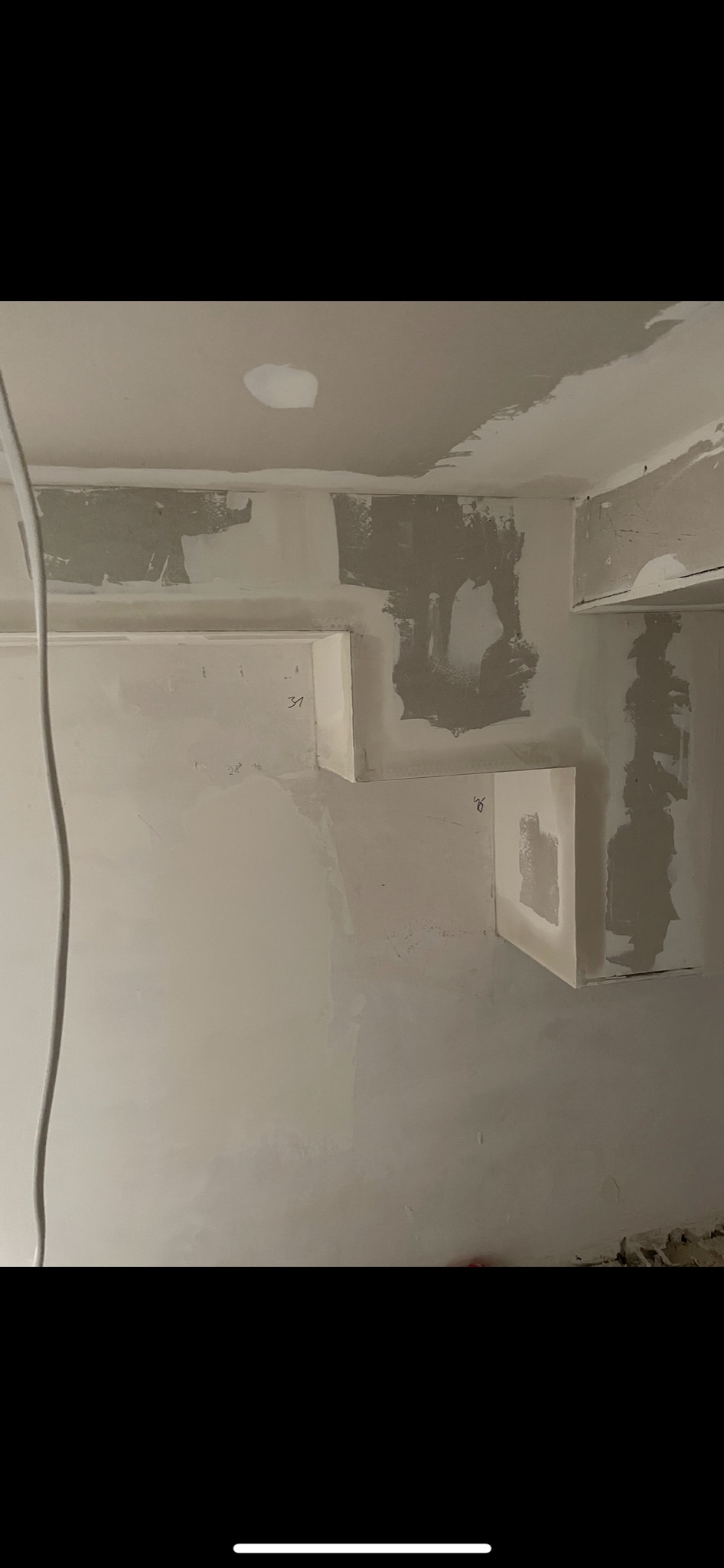 Ściana w trakcie prac wykończeniowych, z widocznymi fragmentami szpachli, niedoskonałościami i zamontowanymi elementami z płyt kartonowo-gipsowych, na których widnieją odręczne notatki.